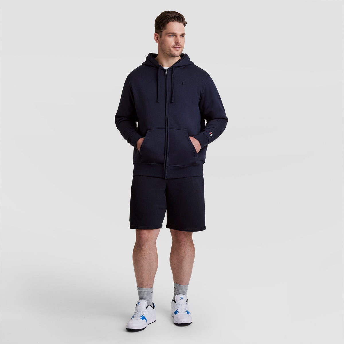 A man wearing a navy Champion Powerblend Full-Zip Hoodie, C Logo - Big & Tall, paired with navy shorts, gray socks, and white sneakers stands with his hands in his hoodie pockets, gazing slightly to the side against a plain light background.