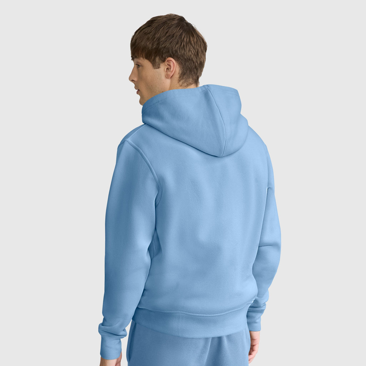 A person with short brown hair is wearing a light blue Champion Powerblend Full-Zip Hoodie, C Logo, paired with matching pants. They are standing and facing away, looking to the left, against a plain light background.