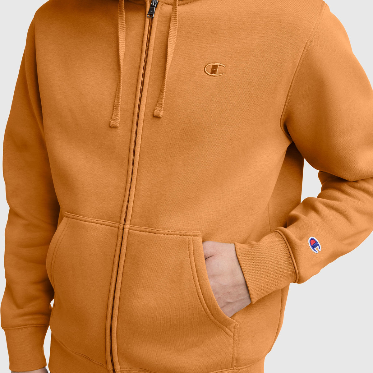A person wearing the Champion Powerblend Full-Zip Hoodie, C Logo in orange has one hand in the front pocket. The iconic Champion logo appears on both the chest and sleeve, showcasing this comfortable men’s full-zip sweatshirt.
