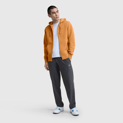 A person wearing the Champion Powerblend Full-Zip Hoodie, C Logo in orange over a white T-shirt, paired with dark gray sweatpants and white sneakers, stands against a plain light gray background, looking slightly to the side.