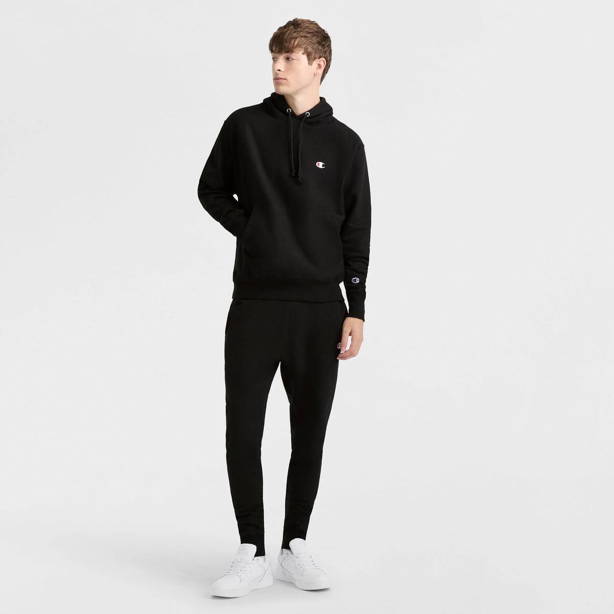 A man in a black Champion Reverse Weave Hoodie, featuring the signature C Logo, stands confidently and showcases this classic style.