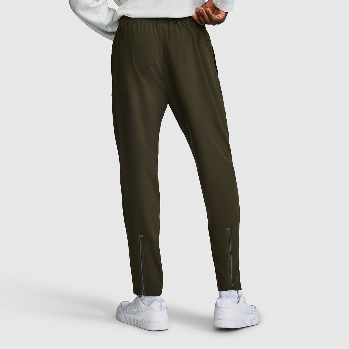A person is shown from the back wearing dark green Champion MVP Pants, Moisture Wicking, C Logo men’s slim-fit sweatpants, paired with a white sweatshirt and white sneakers. The pants feature reflective details near the ankles and are crafted from moisture-wicking fabric for comfort during any activity.