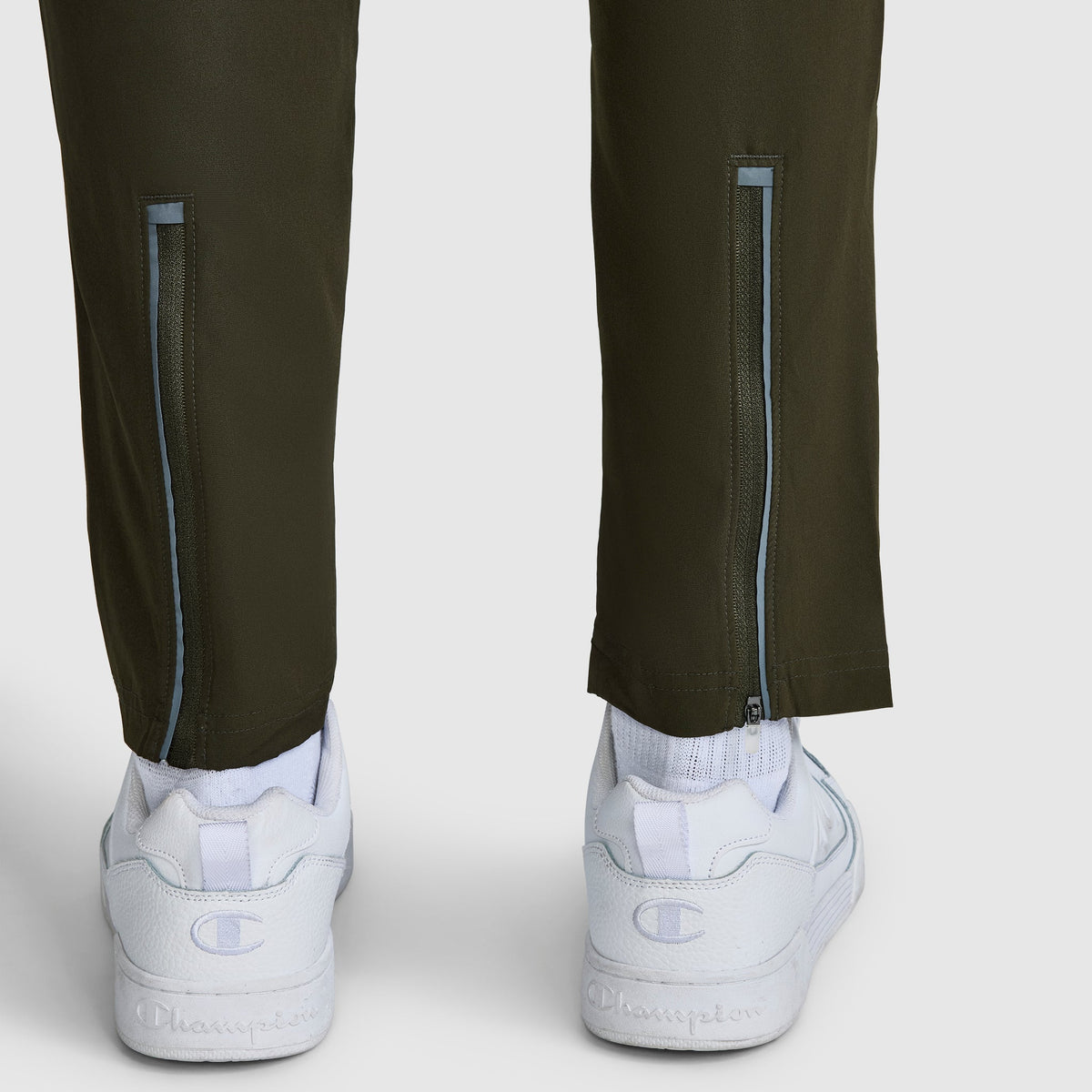 A close-up of the back of a person’s lower legs wearing Champion MVP Pants featuring a slim fit, moisture-wicking fabric, ankle zippers, and the signature C logo, paired with white Champion sneakers.