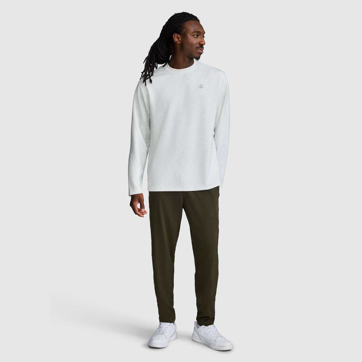 A man stands against a plain light gray background, wearing a long-sleeve white shirt, Champion MVP Pants in dark olive green with ankle zippers and a C logo, which feature moisture-wicking fabric and a slim fit. He completes the look with white sneakers and looks to his left with his left hand relaxed by his side.
