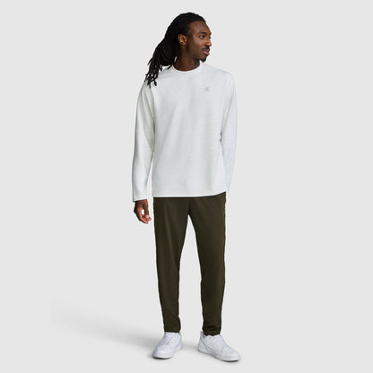 A man stands against a plain light gray background, wearing a long-sleeve white shirt, Champion MVP Pants in dark olive green with ankle zippers and a C logo, which feature moisture-wicking fabric and a slim fit. He completes the look with white sneakers and looks to his left with his left hand relaxed by his side.