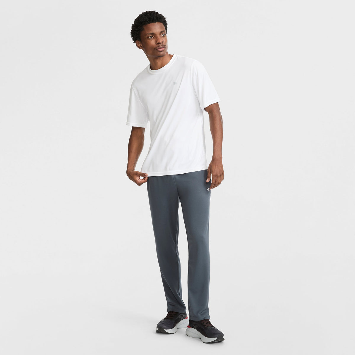 A man stands against a white background wearing a white short-sleeve t-shirt, Champion Sport Sweatpants C Logo with moisture-wicking fabric, and black sneakers. He looks slightly to the side with one hand near his pocket.