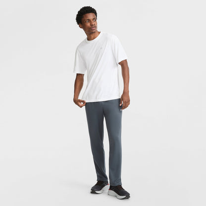 A man stands against a white background wearing a white short-sleeve t-shirt, Champion Sport Sweatpants C Logo with moisture-wicking fabric, and black sneakers. He looks slightly to the side with one hand near his pocket.