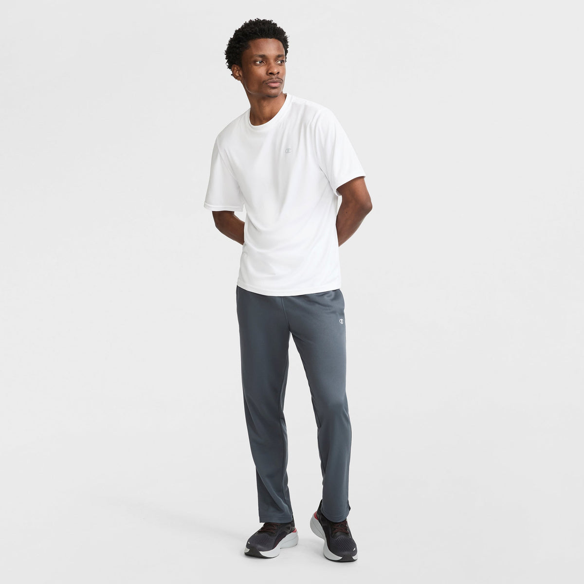 A man stands against a white background wearing a white short-sleeve tee, black sneakers, and Champion Sport Sweatpants with C Logo featuring moisture-wicking fabric for all-day comfort. He looks to the side with his hands behind his back.
