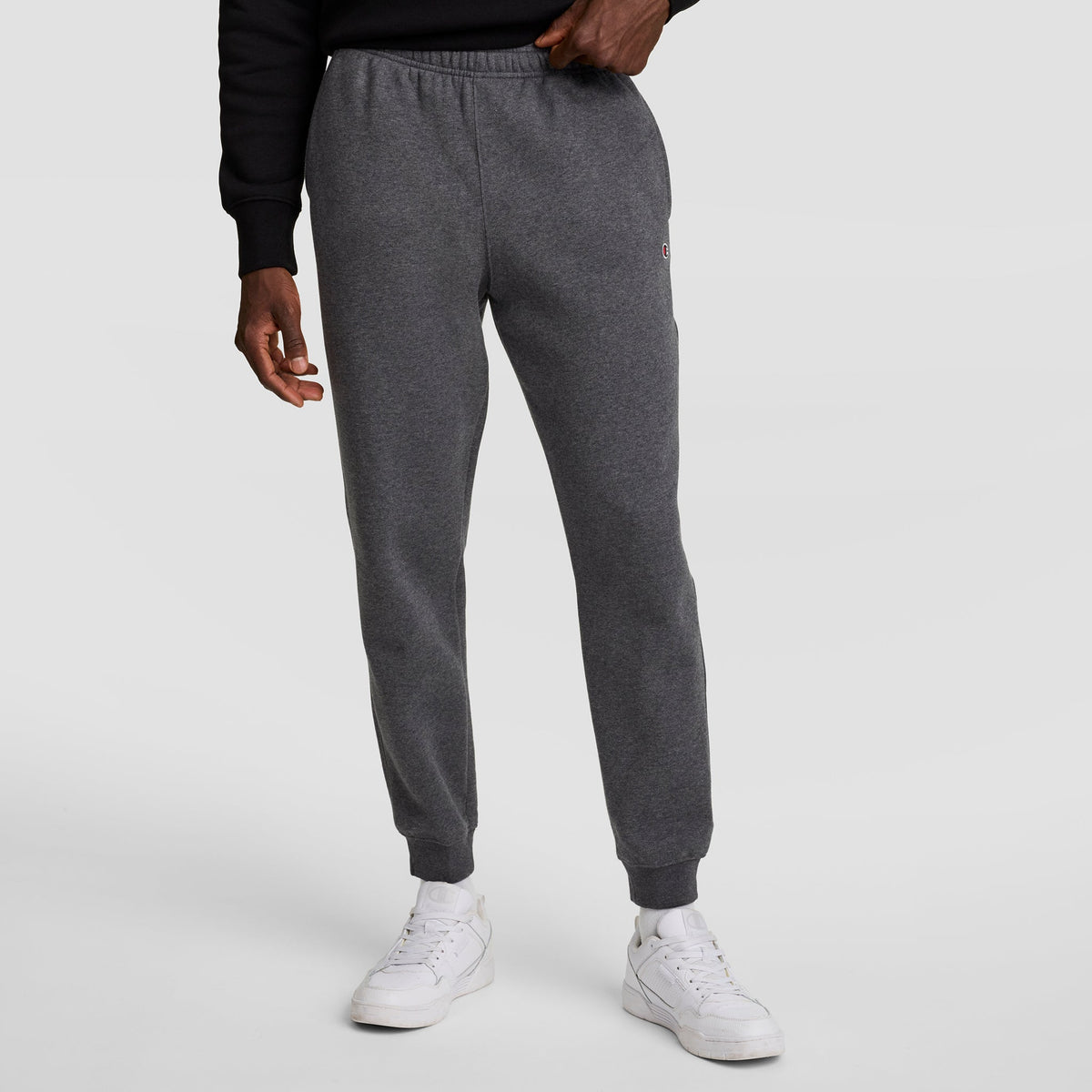 A person wears Champion Powerblend Joggers, C Logo, with a black sweatshirt and white sneakers against a plain light gray background. Only the lower half of the body is visible.
