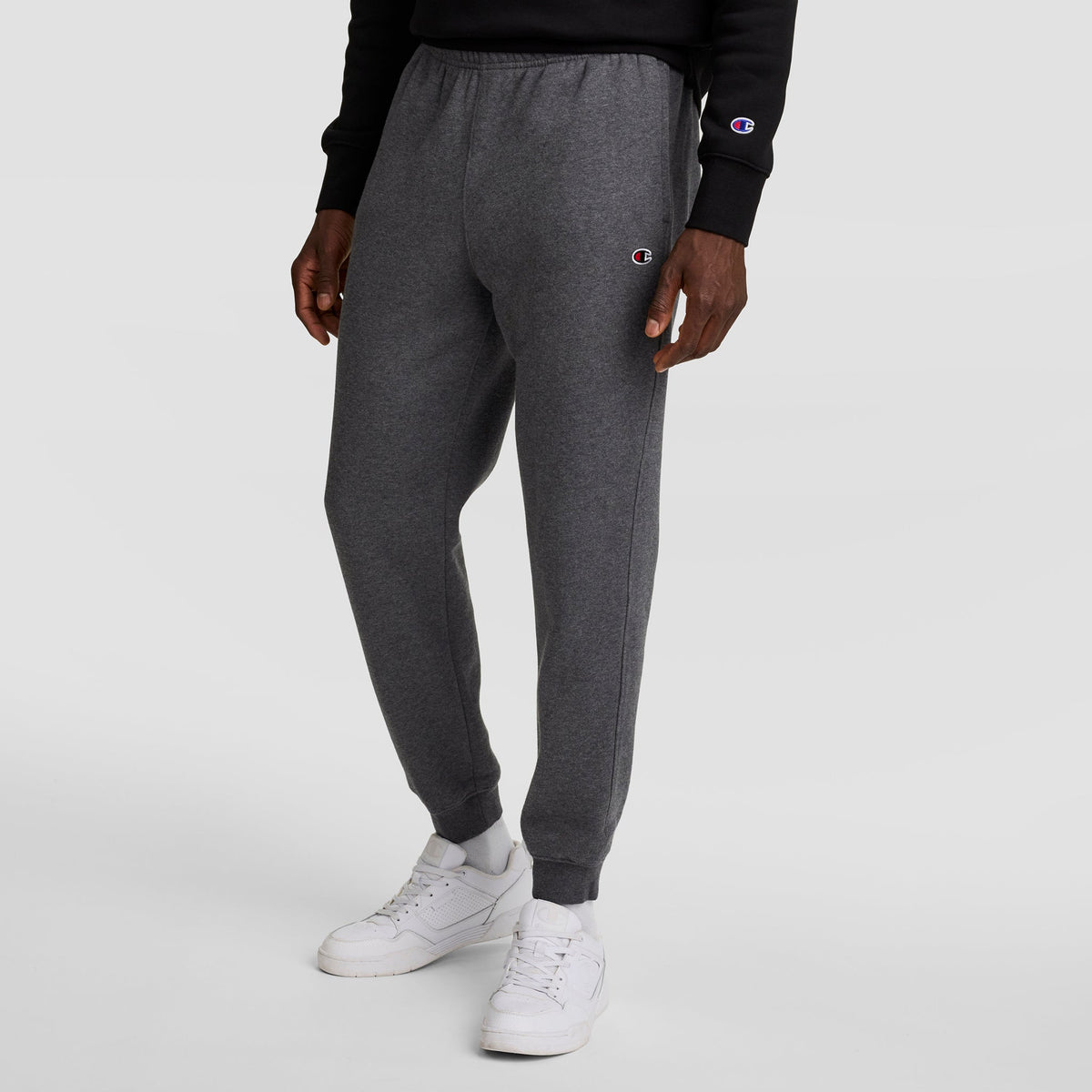 A person wearing Champion Powerblend Joggers, C Logo in dark gray and a black Champion sweatshirt stands against a plain white background. The Champion logo appears on both the joggers and the sweatshirt.