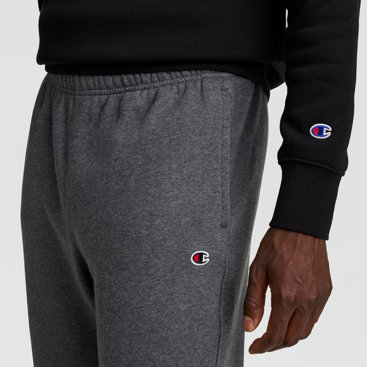 A person wearing dark gray Champion Powerblend Joggers (C Logo), with a black Champion sweatshirt. The visible Champion logos are on the joggers and sweatshirt sleeve. Image shows only the lower torso and hand.