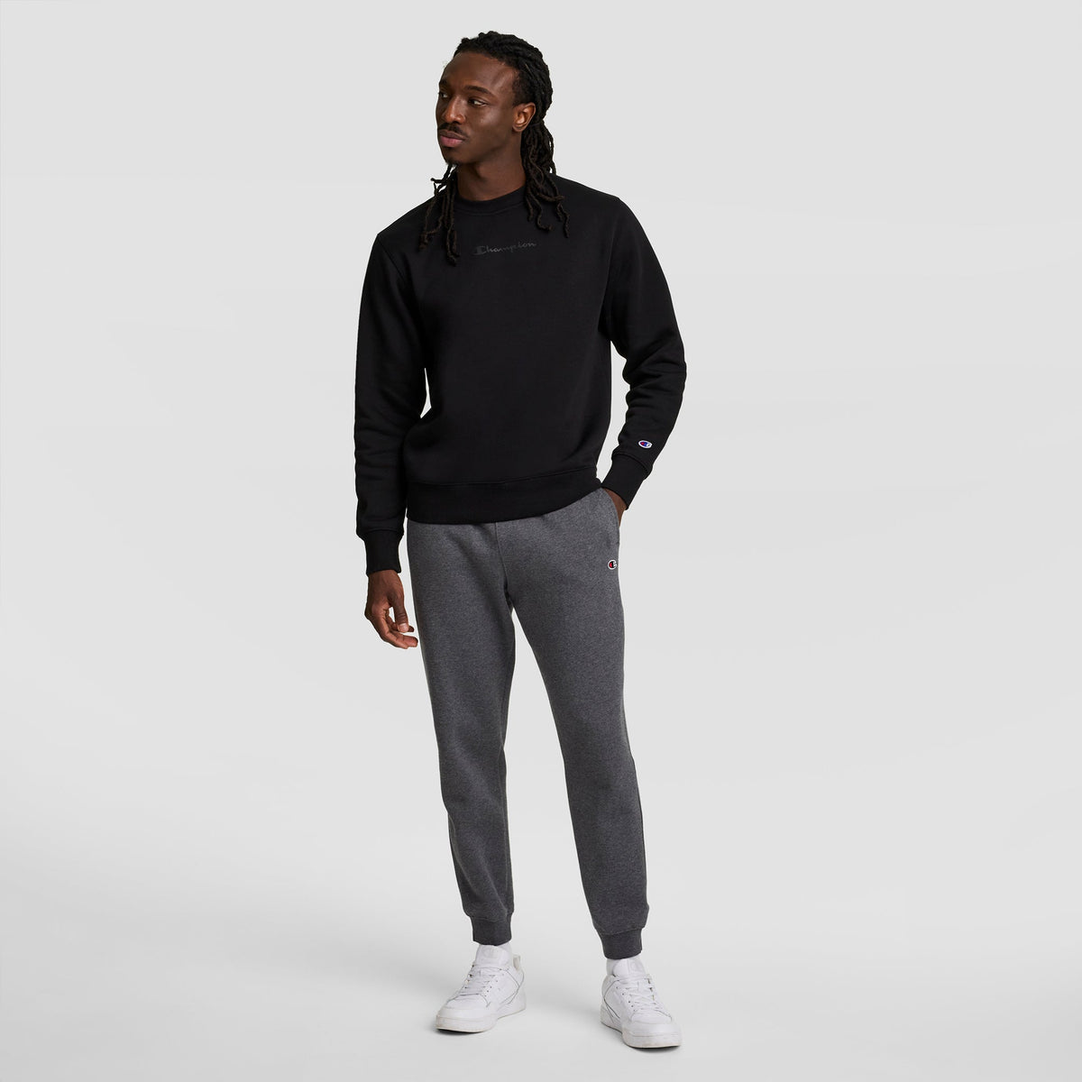 A man with long braided hair wears a black sweatshirt, Champion Powerblend Joggers with the C Logo, and white sneakers, standing against a plain light gray background.