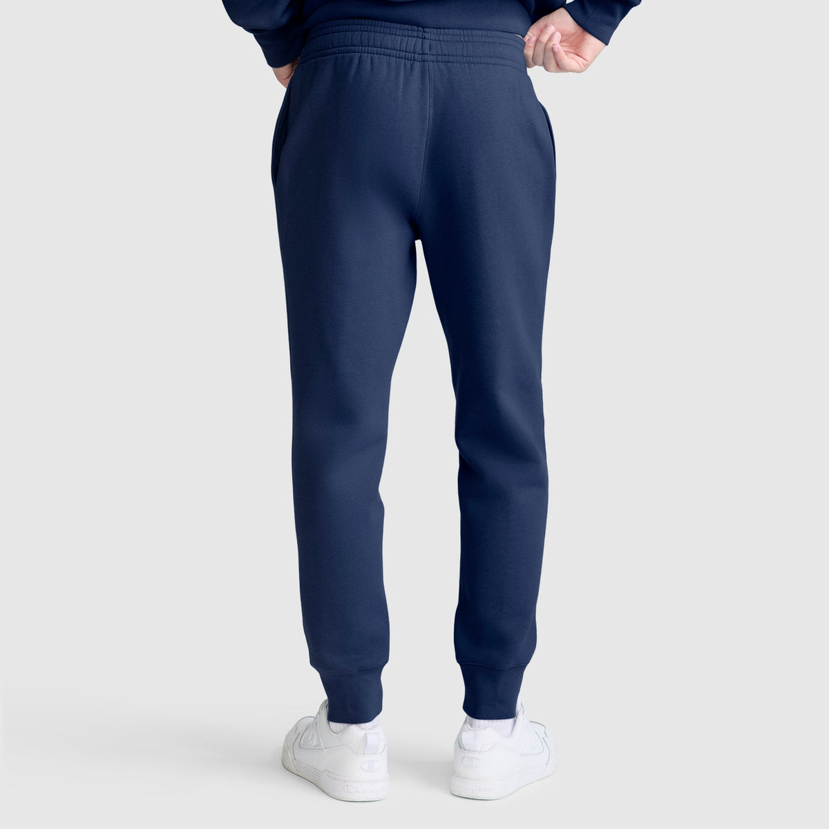 A person stands with their back to the camera, wearing Champion Powerblend Joggers with the C Logo in navy blue and white sneakers against a plain light gray background.