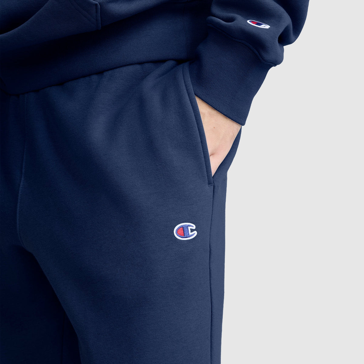 A person wearing a navy blue pair of Champion Powerblend Joggers, C Logo, with their hand in the pocket. The Champion C logo is visible on both the sweatshirt sleeve and the jogger pants, showcasing the classic style of men’s joggers crafted from cozy fleece material.