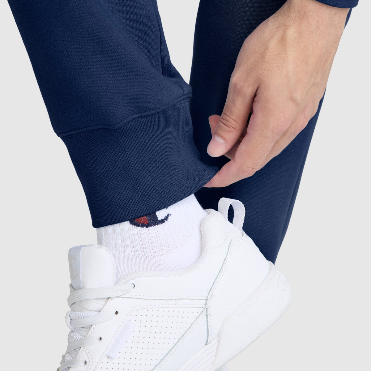 A person wearing Champion Powerblend Joggers featuring the C logo, paired with white logo socks and white athletic sneakers, adjusts the cuff of their pants.