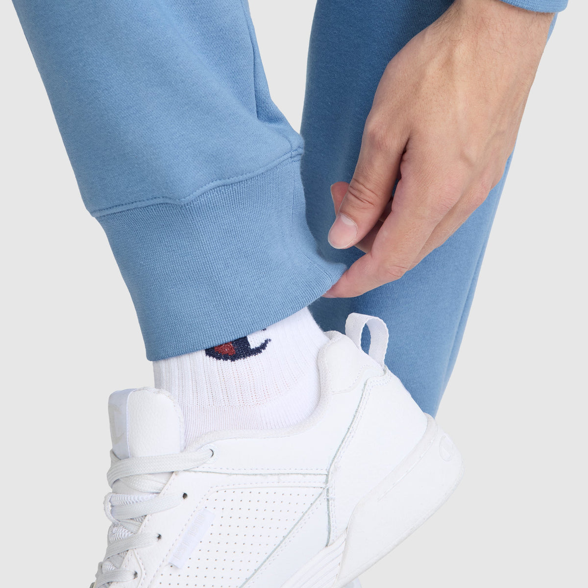 Wearing Champion's Powerblend Joggers with the signature C logo, a person in light blue women's sweatpants adjusts the cuff by their ankle, their white sneakers and white socks highlighting both the comfort and iconic style of these Champion joggers.