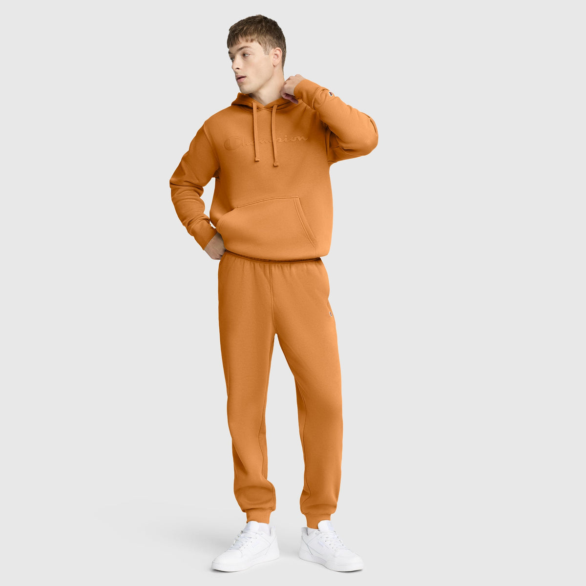 A person stands against a plain background wearing an orange hoodie and Champion Powerblend Joggers, C Logo, paired with white sneakers, looking to the side with one hand on their neck and the other in their pocket.