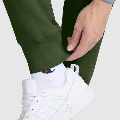 A person wearing green Champion Powerblend Joggers with the C Logo, a blue long sleeve top, white socks, and white sneakers adjusts the cuff of their pants.