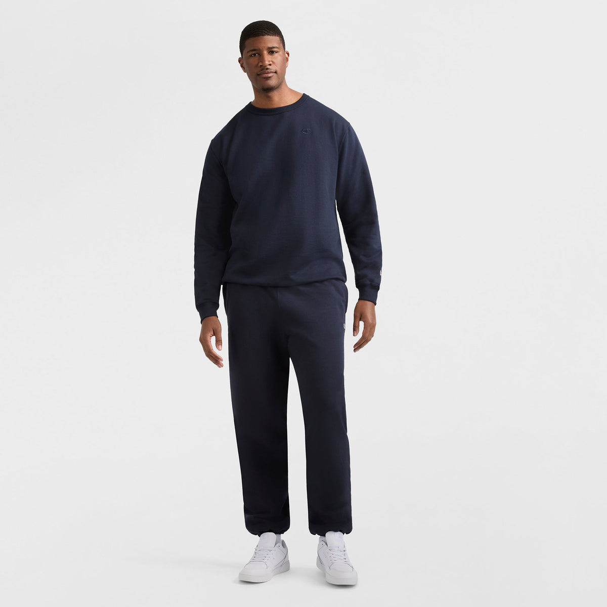 A man stands against a white background, wearing Champion’s Powerblend Sweatpants, Cinched Hem, C Logo - Big & Tall in dark navy with white sneakers. He faces forward with a relaxed posture, showcasing these athletic sweatpants.