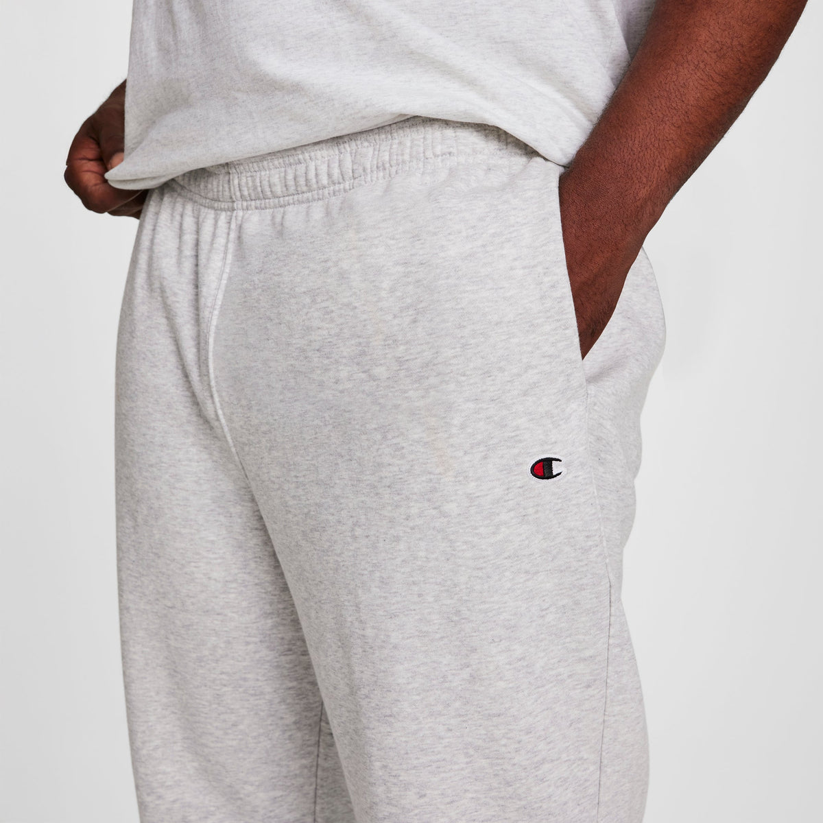 A person wears light gray Champion Powerblend Sweatpants, Cinched Hem, C Logo - Big & Tall, with a matching t-shirt and one hand in their pocket. The Champion logo is visible on the sweatpants' cinched hem.
