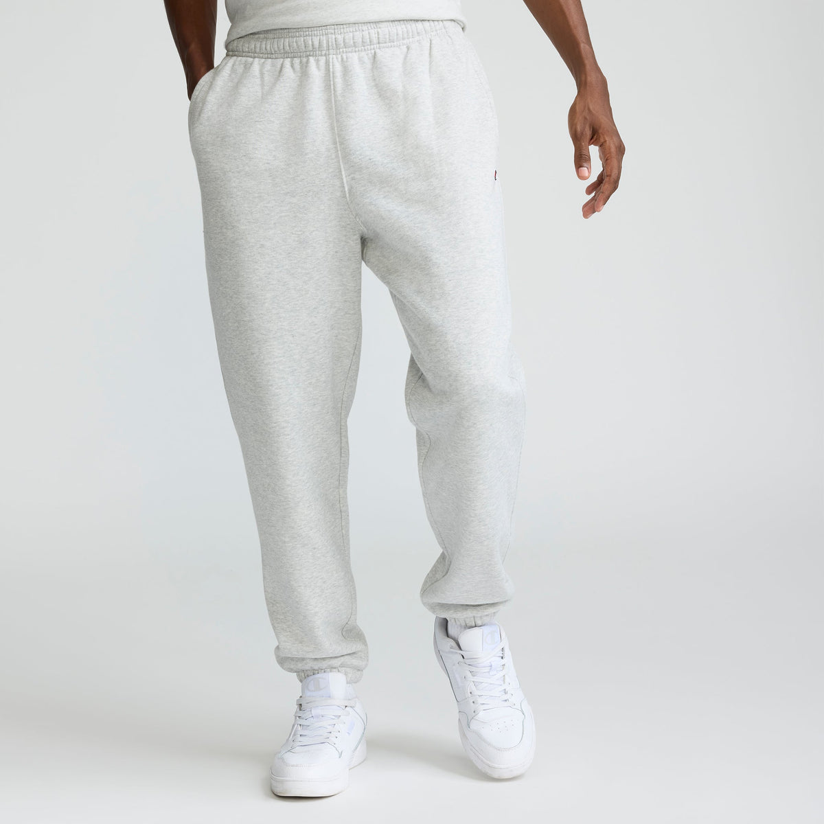 A person wearing Champion Powerblend Sweatpants, Cinched Hem, C Logo - Big & Tall in light gray and white sneakers stands with hands in pockets against a plain white background. The upper body is mostly out of frame.