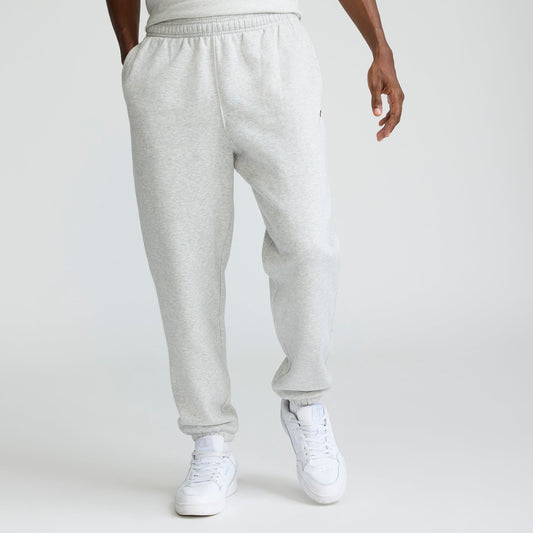 A person wearing Champion Powerblend Sweatpants, Cinched Hem, C Logo - Big & Tall in light gray and white sneakers stands with hands in pockets against a plain white background. The upper body is mostly out of frame.