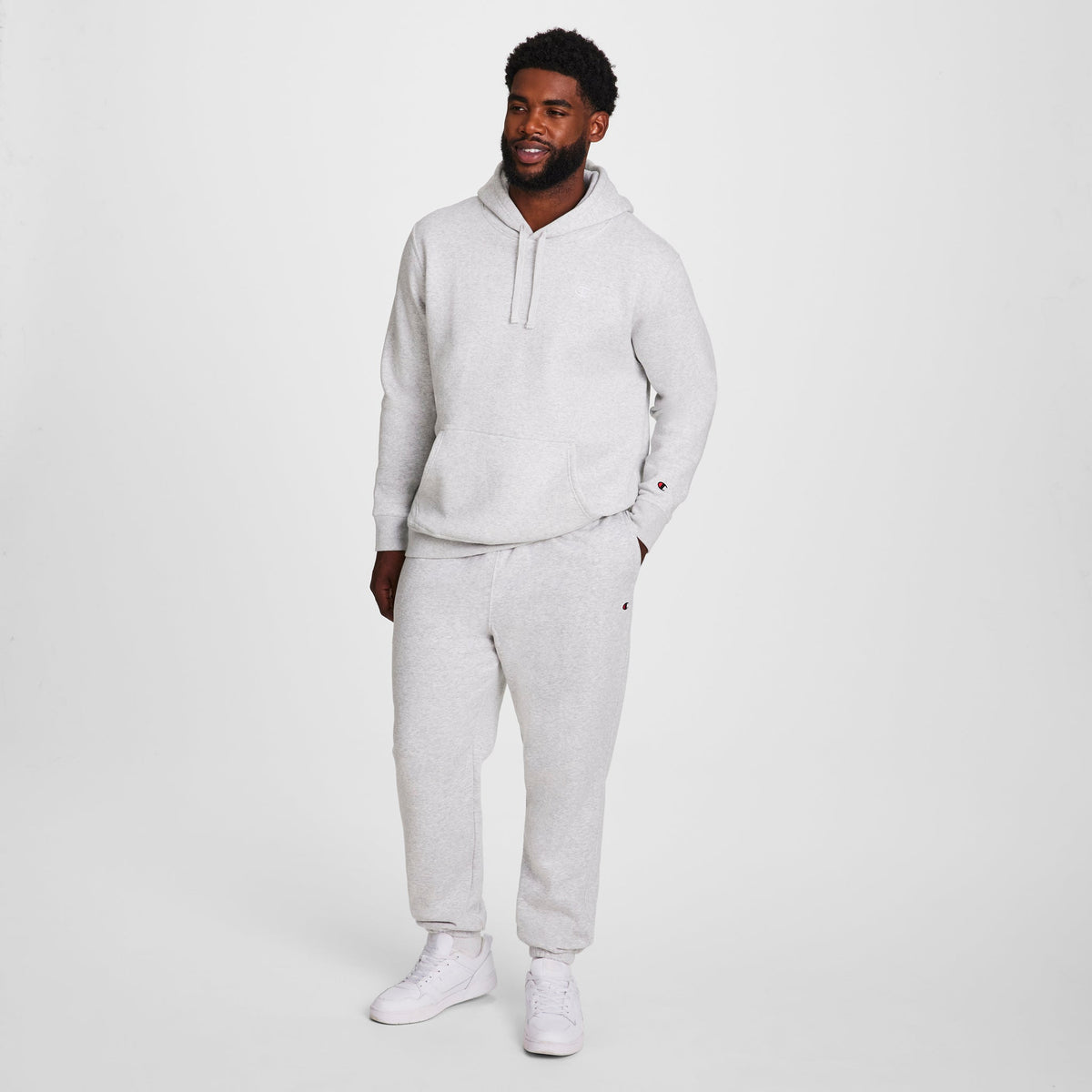 A man smiles with his hands by his sides, wearing Champion Powerblend Sweatpants, Cinched Hem, C Logo - Big & Tall in light gray and matching sweatshirt, white sneakers, standing against a plain white background.