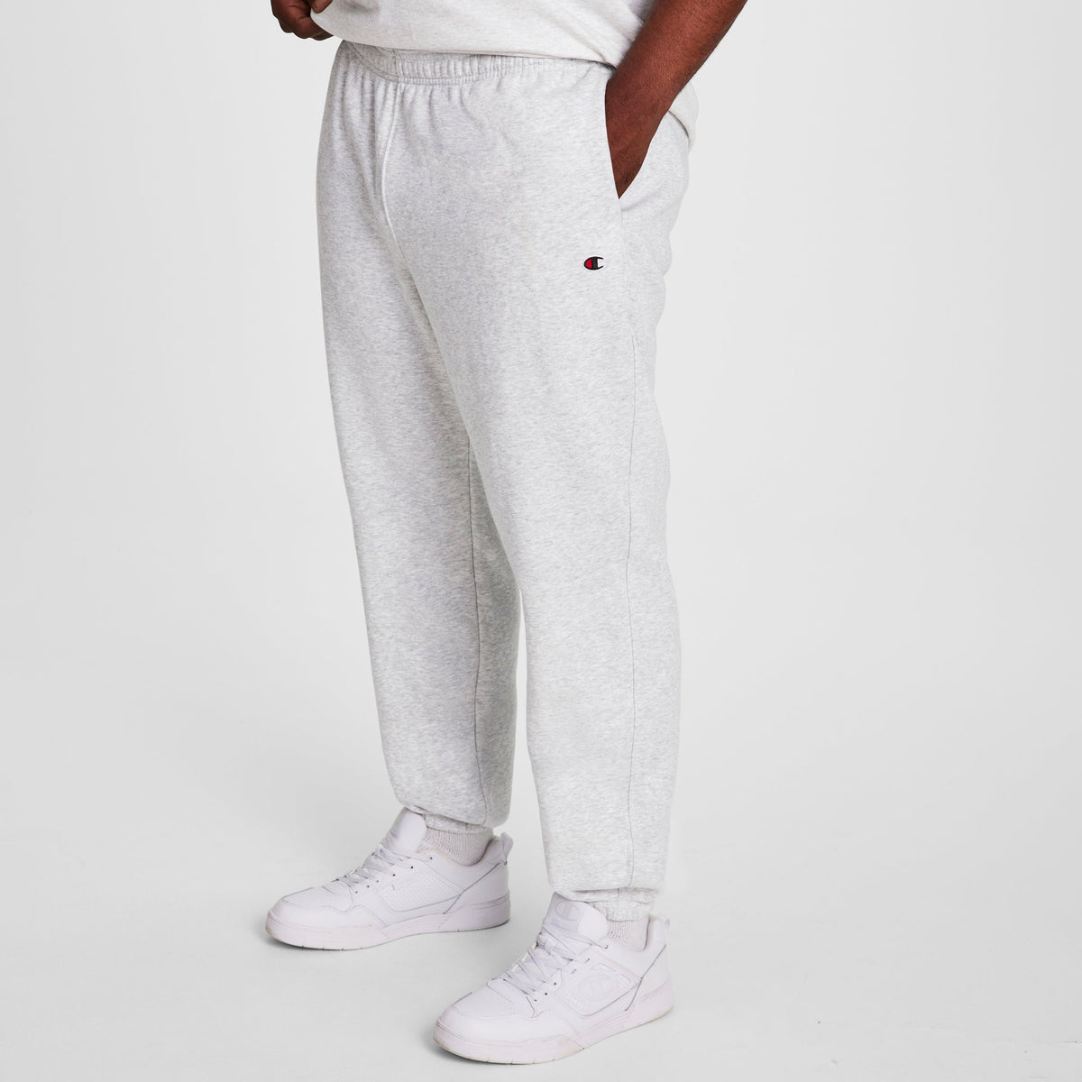 A person models Champion Powerblend Sweatpants, Cinched Hem, C Logo - Big & Tall in light gray, paired with white sneakers. Only their lower body is shown against a white background.