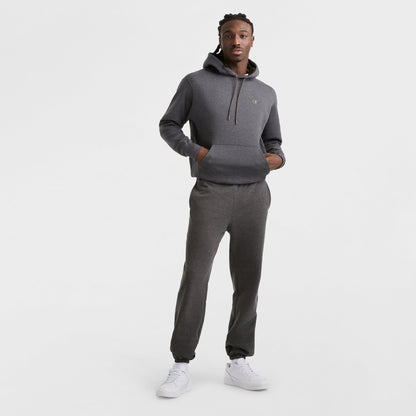 A man stands against a white background wearing Champion Powerblend Sweatpants, Cinched Bottom, C Logo in dark gray with matching hoodie and white sneakers, hands in the hoodie pocket, looking at the camera.