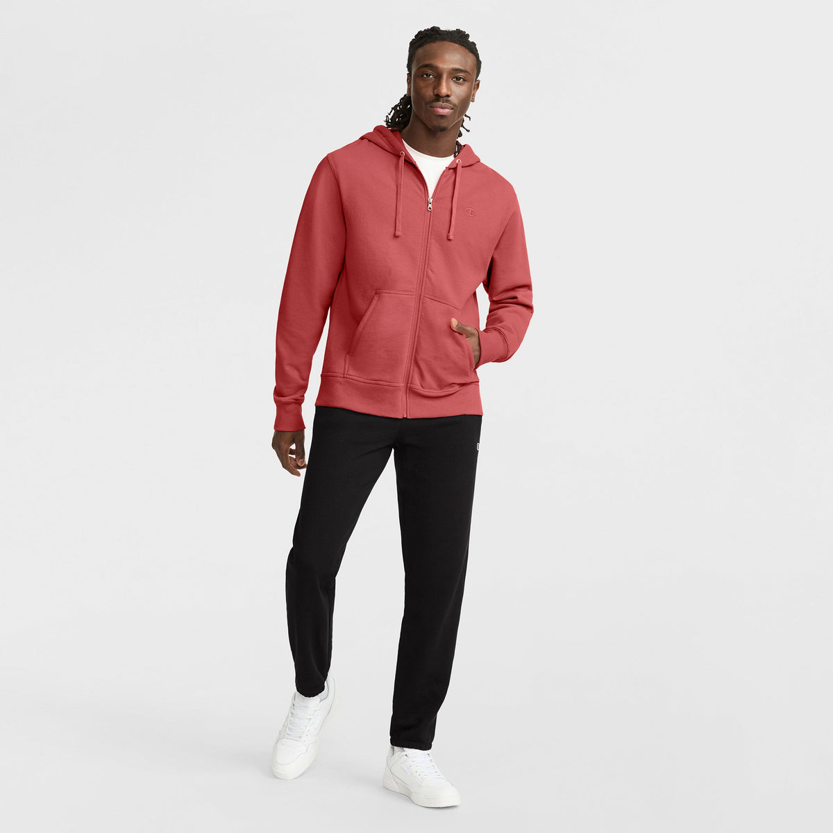 A man stands against a white background wearing a red zip-up hoodie, Champion Powerblend Sweatpants with Cinched Bottom and C Logo in black, and white sneakers. He has long twisted hair and looks slightly to the side with a relaxed expression.