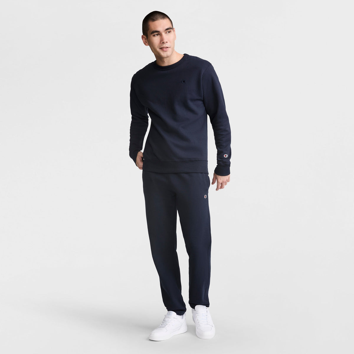 A man stands against a plain white background wearing Champion Powerblend Sweatpants, Cinched Bottom, C Logo in navy blue with a matching sweatshirt and white sneakers, looking slightly to the side with one hand in his pocket.