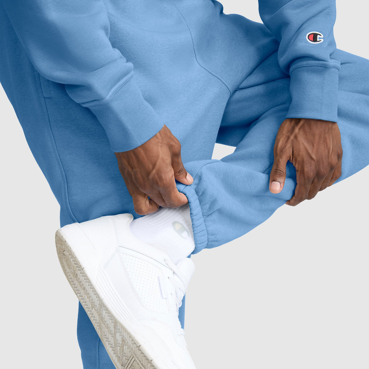 A person wearing light blue Champion Powerblend Sweatpants, Cinched Bottom, C Logo, along with a matching sweatshirt featuring the Champion logo, adjusts the pant leg to reveal white socks and white sneakers against a light background.