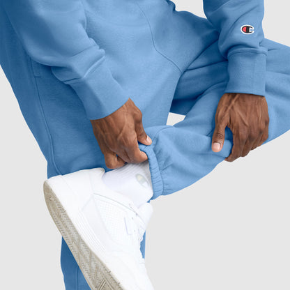 A person wearing light blue Champion Powerblend Sweatpants, Cinched Bottom, C Logo, along with a matching sweatshirt featuring the Champion logo, adjusts the pant leg to reveal white socks and white sneakers against a light background.