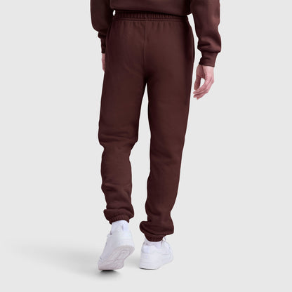 A person wearing Champion Powerblend Sweatpants with a cinched bottom and prominent C logo is shown from the back, standing against a plain light gray background. The brown fleece pants are paired with white sneakers.