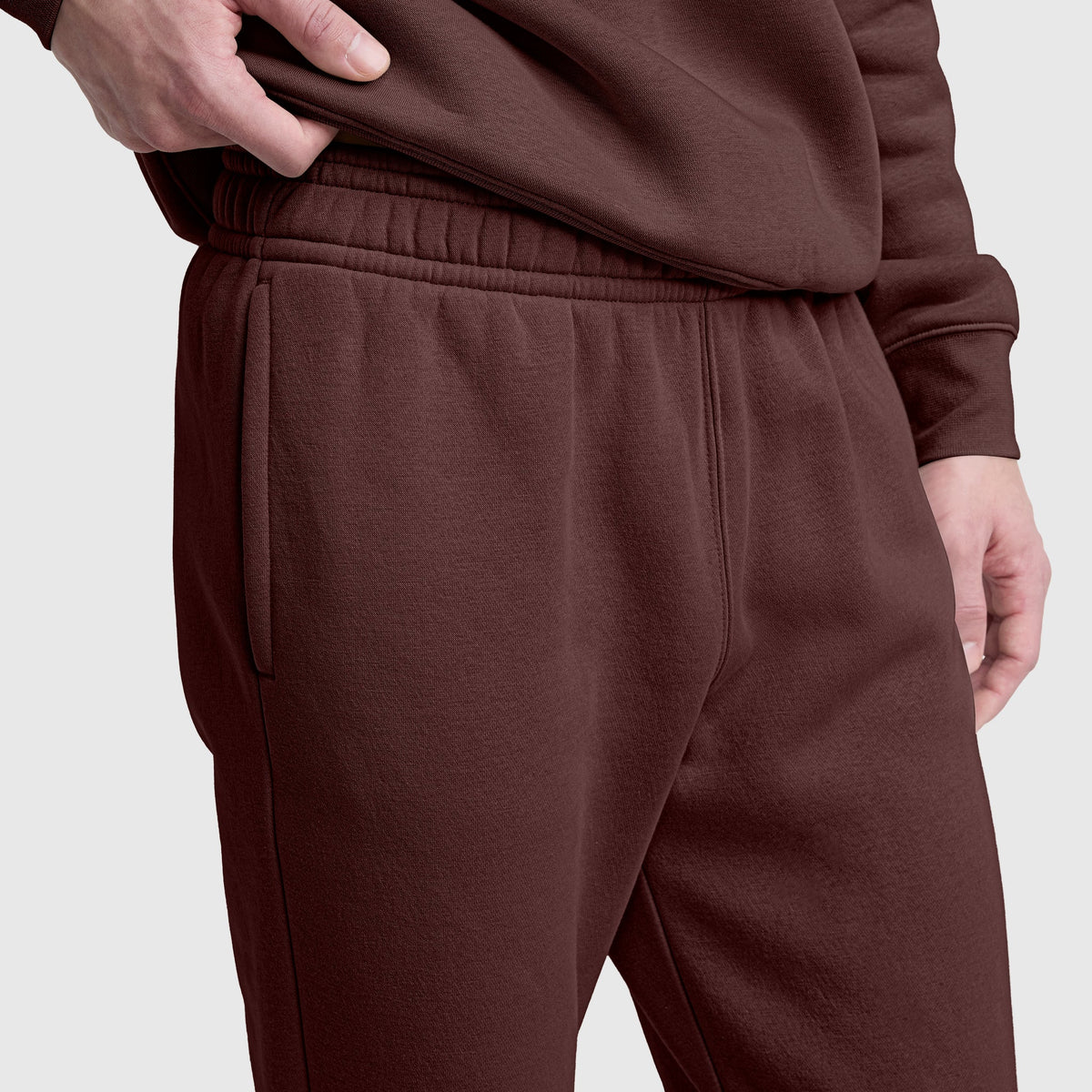 A person stands against a plain light gray background wearing matching brown Champion Powerblend Sweatpants with a cinched bottom and C logo—one hand in their pocket, the other resting by their side.