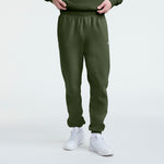 A person is wearing Champion Powerblend Sweatpants with a cinched bottom and C logo in olive green, paired with white sneakers, standing against a plain white background. Only the lower half of the body is visible.