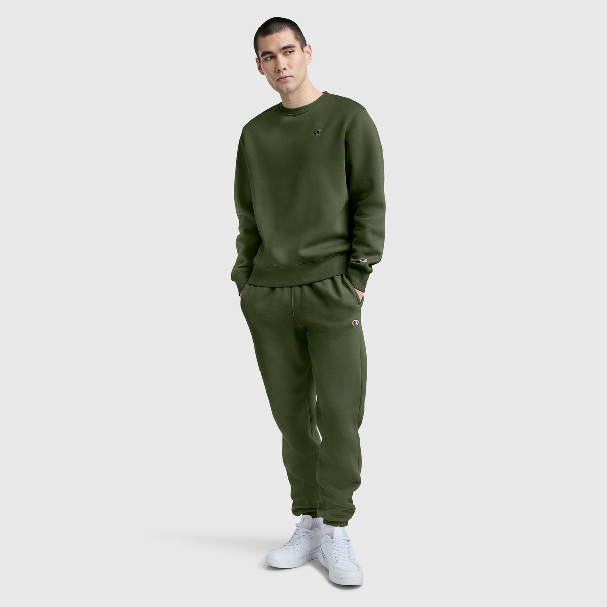 A person stands against a plain light gray background, wearing a matching olive green sweatshirt and Champion Powerblend Sweatpants with Cinched Bottom and C Logo, paired with white sneakers.