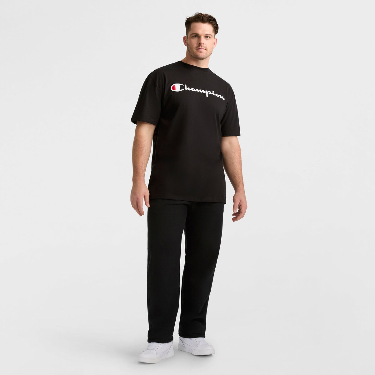 A man stands against a plain white background, wearing a black Champion t-shirt, white sneakers, and Champion Powerblend Sweatpants, Open Hem, C Logo - Big & Tall. He faces forward with a relaxed posture.