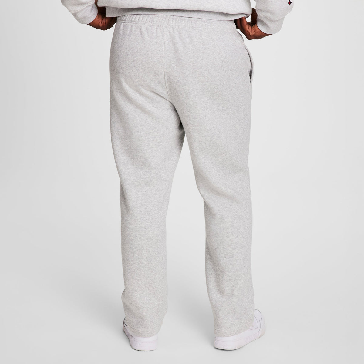 A person stands facing away against a white background, wearing Champion Powerblend Sweatpants, Open Hem, C Logo - Big & Tall in light gray with a matching sweatshirt and white sneakers. Hands are on hips.