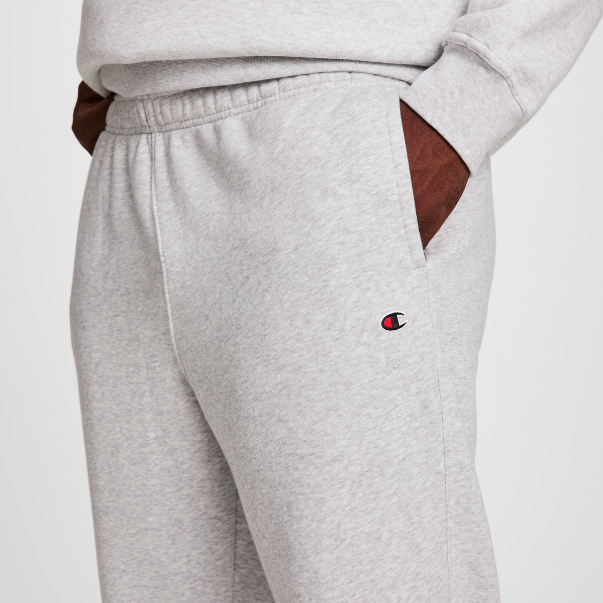 A person wears Champion Powerblend Sweatpants, Open Hem, C Logo - Big & Tall in light gray with a matching sweatshirt. The Champion logo appears near the left pocket. Only the torso and upper legs are visible, hands in pockets.