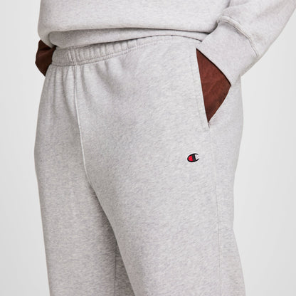 A person wears Champion Powerblend Sweatpants, Open Hem, C Logo - Big & Tall in light gray with a matching sweatshirt. The Champion logo appears near the left pocket. Only the torso and upper legs are visible, hands in pockets.