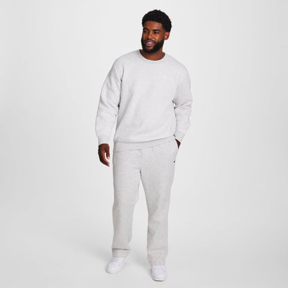 A man smiles in a Champion Powerblend Sweatpants, Open Hem, C Logo - Big & Tall and a light gray cotton blend sweatshirt with white sneakers, standing against a plain white background.