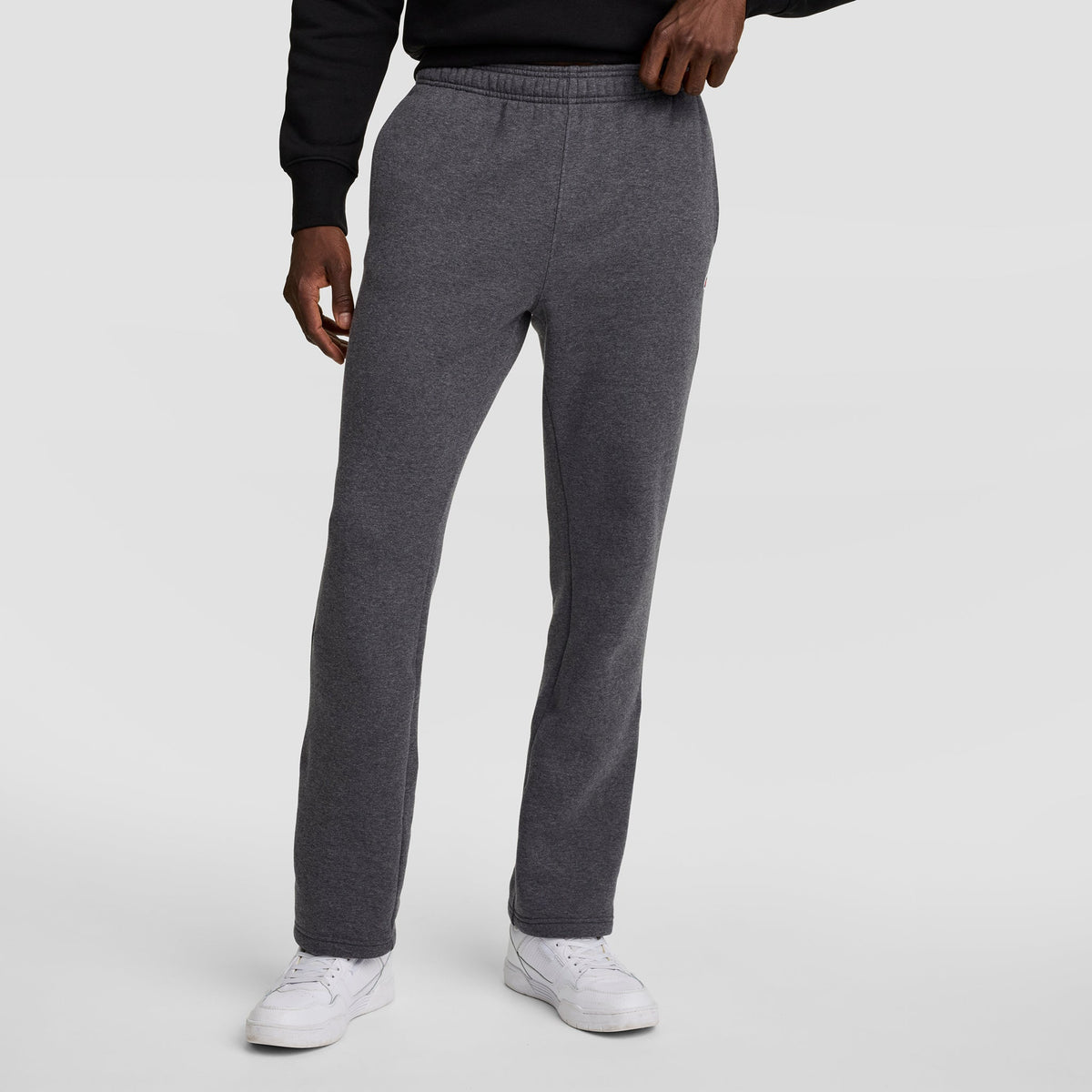 A person wearing Champion Powerblend Sweatpants, Open Hem, C Logo in charcoal gray stands against a plain light gray background, paired with a black sweatshirt and white sneakers, left hand resting on their waistband.