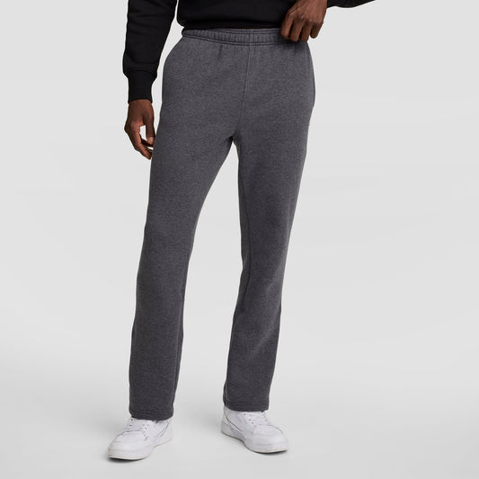 A person wearing Champion Powerblend Sweatpants, Open Hem, C Logo in charcoal gray stands against a plain light gray background, paired with a black sweatshirt and white sneakers, left hand resting on their waistband.