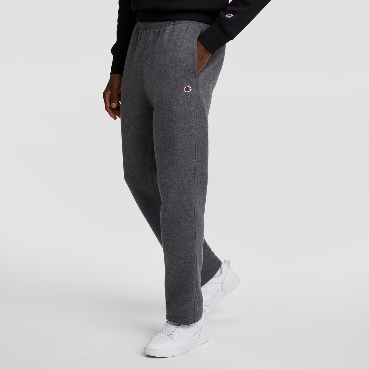 A person in dark gray Champion Powerblend Sweatpants (Open Hem, C Logo), a black sweatshirt, and white sneakers stands against a light gray background. The Champion logo is visible on the sweatpants and sweatshirt sleeve.