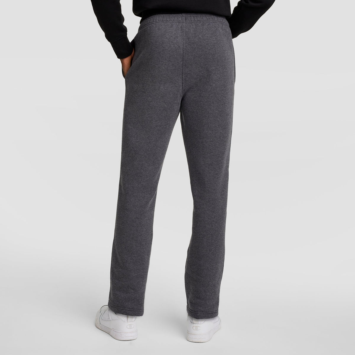 Someone is shown from the back with hands in pockets, wearing dark gray Champion Powerblend Sweatpants, Open Hem, C Logo, a black top, and white sneakers against a plain light background.