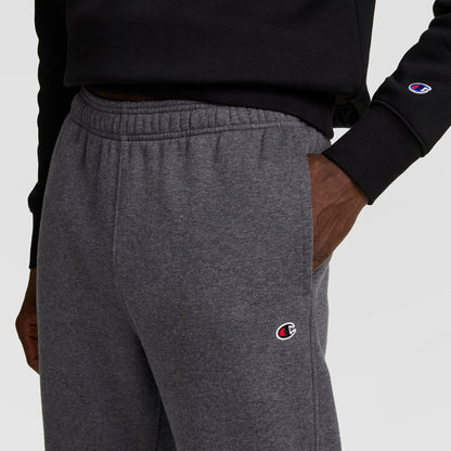 A person wears a black sweatshirt and gray Champion Powerblend Sweatpants, Open Hem, C Logo, with one hand in their pocket. Both the sweatshirt cuff and the sweatpants show the Champion logo.