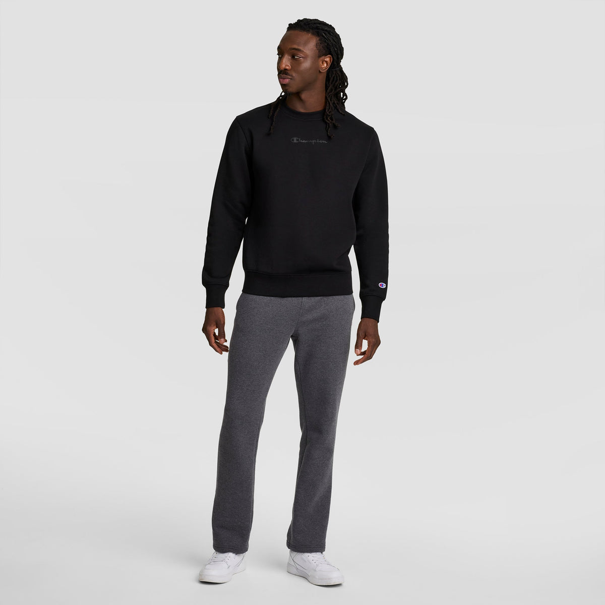 A man faces slightly left, wearing a black sweatshirt, Champion Powerblend Sweatpants with an open hem and C Logo, plus white sneakers, against a plain light gray background.