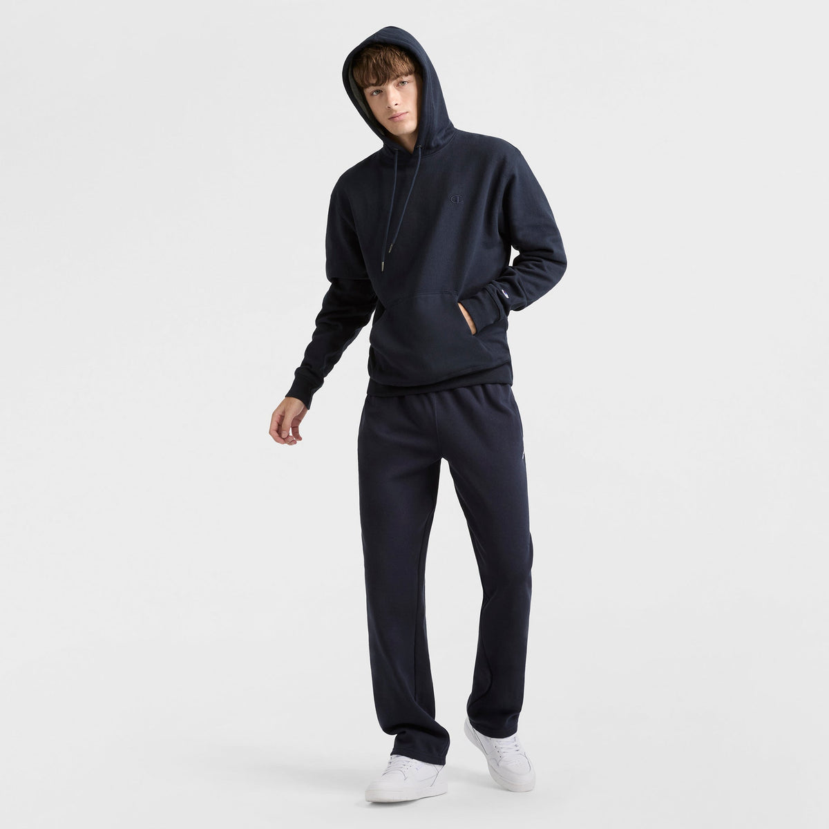A person wearing Champion Powerblend Sweatpants, Open Hem, C Logo, and a dark hooded sweatshirt with white sneakers stands against a plain white background.