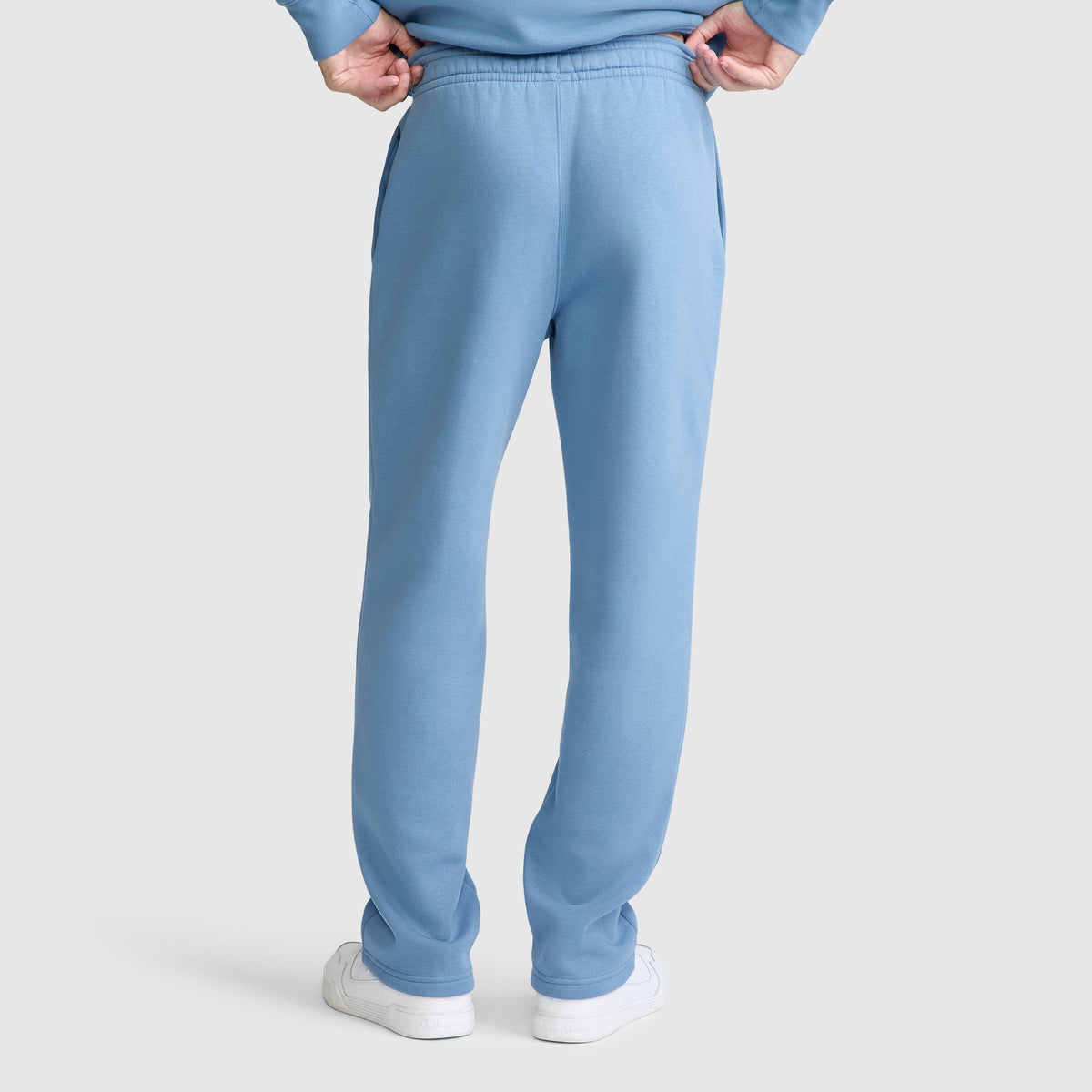 A person is seen from behind, standing with hands on hips and wearing Champion Powerblend Sweatpants, Relaxed Bottom, C Logo in light blue along with white sneakers. The image highlights the back view of the pants against a plain light background.